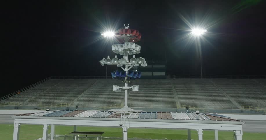 An elevated night view shows a tall rotating speaker tower with a crown like top and bright spotlights over a small structure with empty stadium bleachers and a race track visible in the dark background