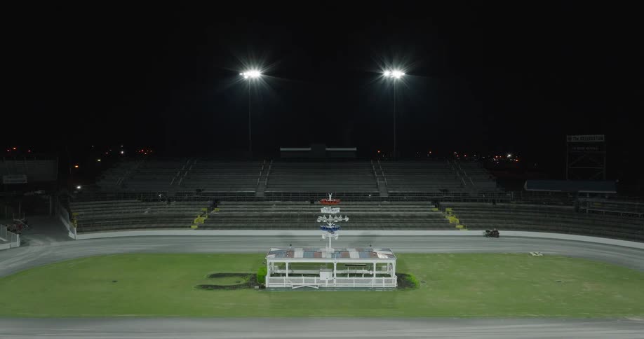 A large empty racetrack stadium at night two bright floodlights illuminate the track and surrounding tiers of vacant seating a small white building with a central mast stands in the middle of the green infield