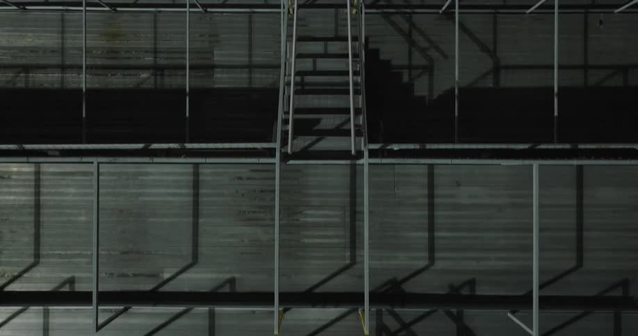 An aerial shot looks straight down onto a long tiered stadium seating area with metal staircases and handrails casting diagonal shadows across the dark surface
