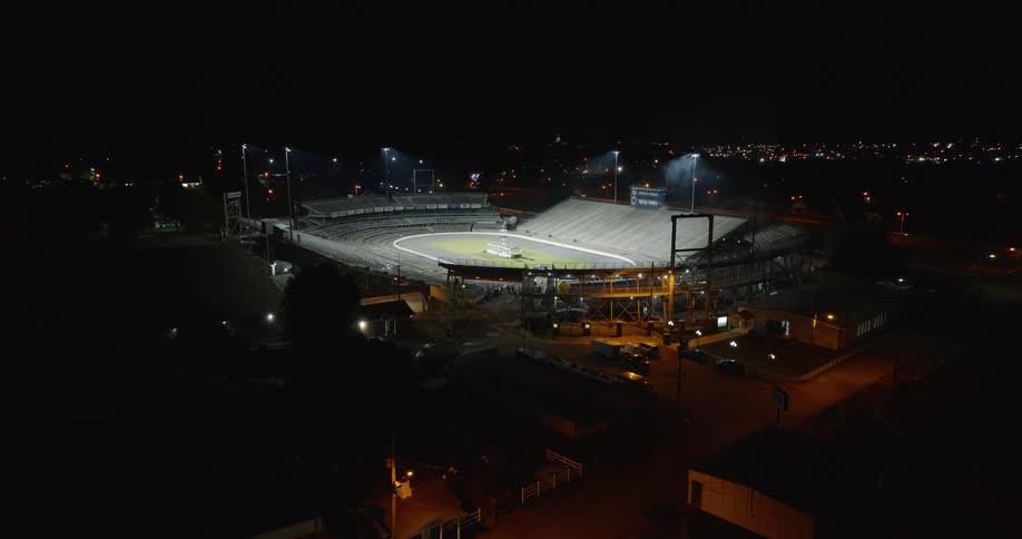 The video is a high wideangle aerial shot of a speedway or dirt track stadium lit up at night the surrounding area is dark with distant scattered city lights in the background