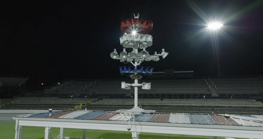 A tall siren on a pole above a low structure with a striped roof the device is centered in the foreground while rows of empty stadium bleachers stretch out into the dark background and floodlights illuminate the scene
