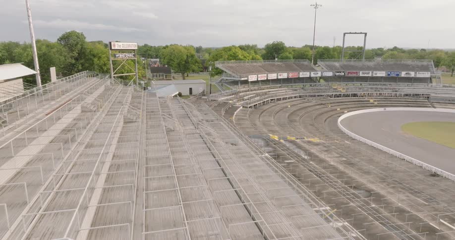 An elevated camera shot slowly pans right over wide empty stadium grandstands and rows of seating that curve around a section of an oval racetrack under a cloudy sky