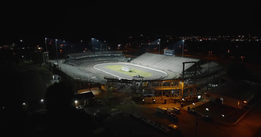 The video is a high wideangle aerial shot of a large welllit speedway or dirt track stadium at night the stadium itself is bright standing out against the dark surrounding area with scattered city lights in the distance