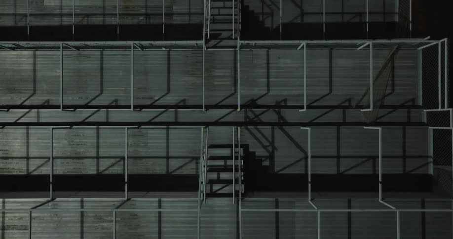 An abstract overhead drone shot shows the geometric pattern of empty tiered outdoor seating with metal railings and shadows cast by the bright sun as the camera pushes in and rotates over the dark seating rows