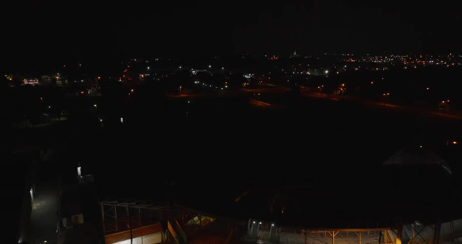 An elevated sweeping aerial shot captures a large empty stadium with bleachers and a race track where a small utility vehicle is visible on the grass infield all illuminated by bright overhead lights against the dark surrounding city night