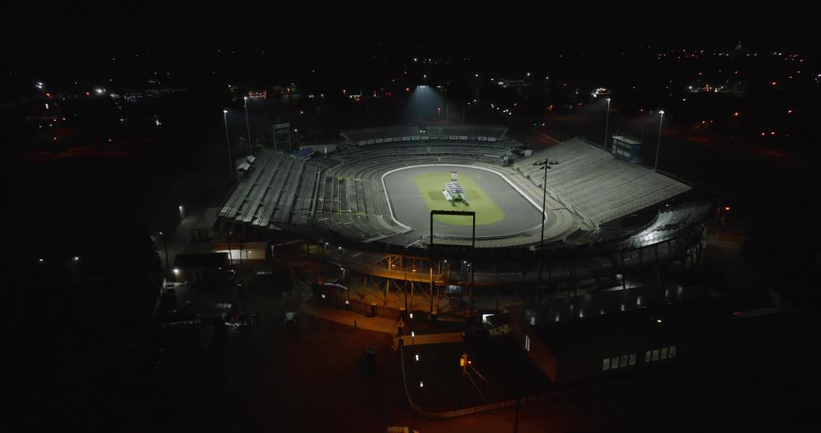 An overhead shot shows a brightly lit oval racetrack at night a striped vehicle moves across the infield grass and track surface tiers of dark empty spectator seating surround the circuit and the camera reveals the wider stadium area