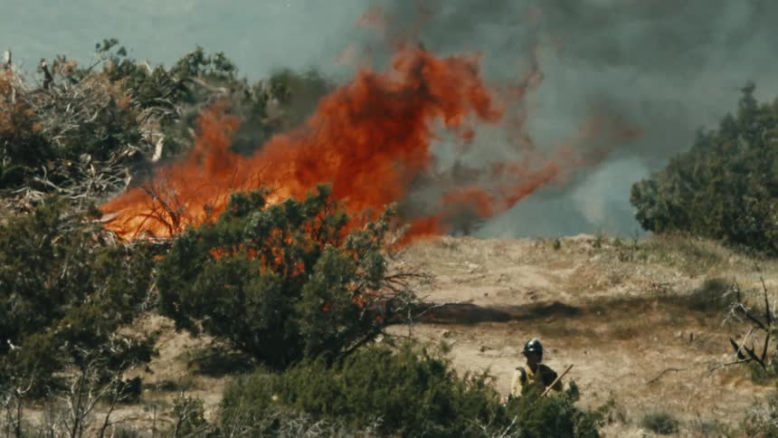 The video is a medium shot of a fire burning in a dry brushy landscape producing bright orange flames and a plume of dark gray and orange smoke in the lower right corner a figure wearing a helmet and a protective jacket is partially visible i