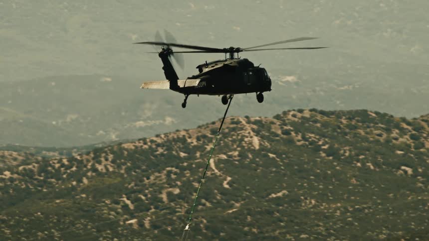 A helicopter flies over a field at midday dragging a long rope below