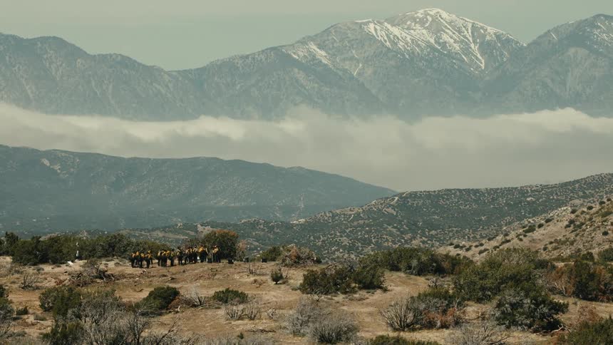 The video shows a wide shot of a rugged dry landscape with a group of people possibly firefighters in yellow gear gathered in the middle ground the background is dominated by a majestic chain of tall mountains with the highest peak capped i