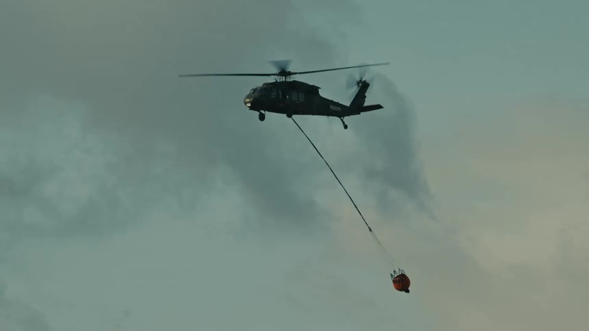 A long view of a dark military helicopter silhouetted against a pale cloudy sky as it carries a bright orange water drop bucket on a long cable