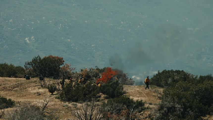A high angle wide view shows several firefighters in yellow work clothes working around a controlled burn in a dry brush covered mountain landscape under a slightly hazy sky