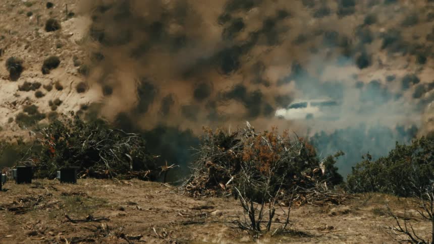 Dark smoke rising from a fire in a gully near a yard partially obscures a passing vehicle