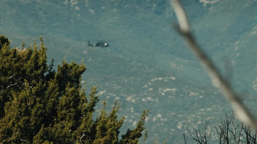 A helicopter flies with green trees on a mountain slope in the background