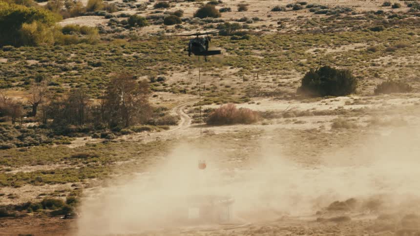 A military helicopter flies over a dry brush filled landscape carrying a red bucket on a long cable while kicking up a large amount of dust below
