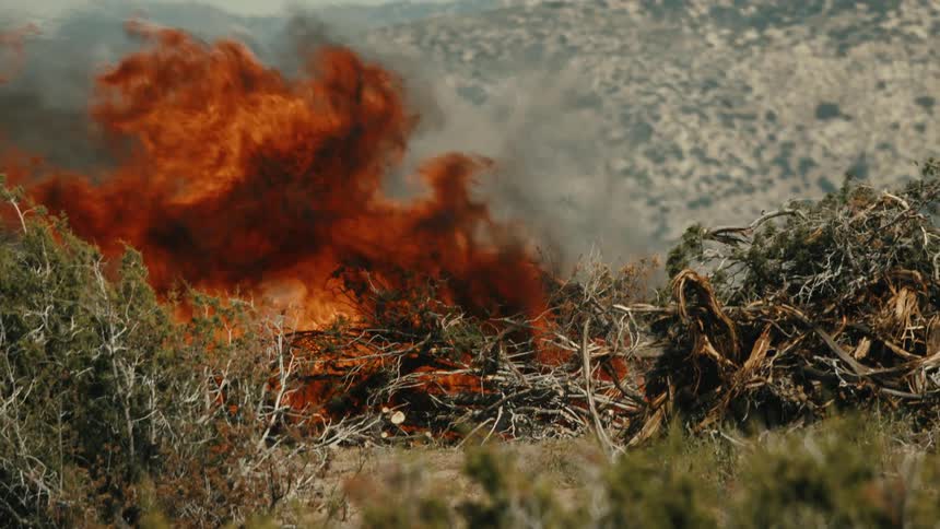 The video shows a large pile of dry brush and dead wood burning fiercely in the center of a scrubcovered mountainous landscape the huge orange flames and thick plume of reddishbrown smoke dominate the foreground against the light brown dry hi