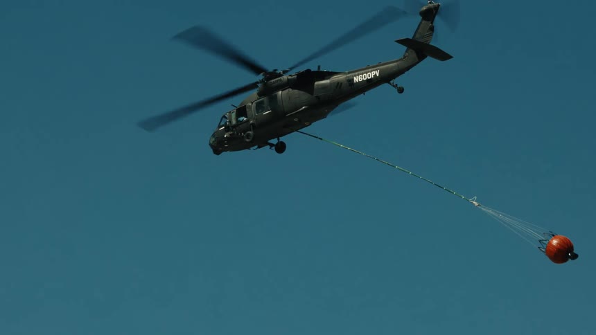 A helicopter carries an orange water bucket across a clear blue sky