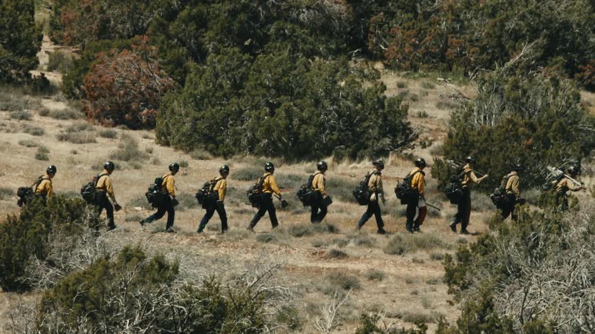 A group of wildland firefighters walk through dry brush carrying packs