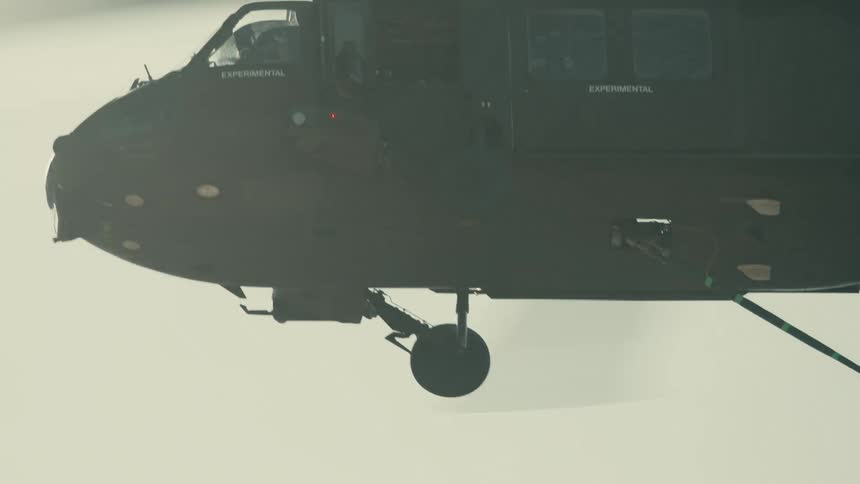 The video shows a darkcolored military or searchandrescue helicopter flying close to the ground with dust or snow swirling up beneath its body the camera is close to the aircraft and the background is almost entirely blurred out by the bright