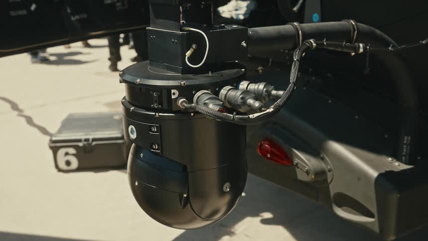The video is an extreme closeup shot of a complex black camera or sensor gimbal mounted to the exterior of an aircraft or drone the device is highly detailed showing cables and connectors against a bright sunlit background