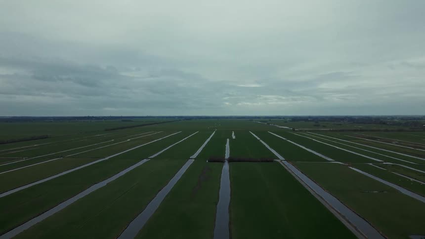 The video is an aerial drone shot flying low over a vast expanse of flat bright green agricultural land that is clearly divided into long narrow plots by horizontal and vertical water channels or ditches under an overcast sky this landscape su