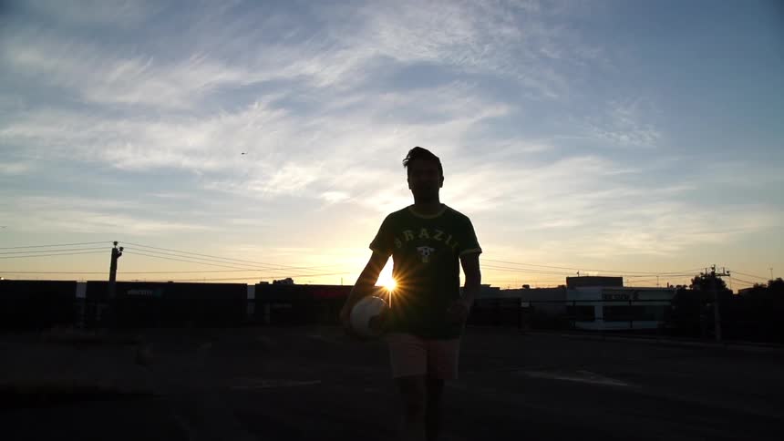 A man walks holding a soccer ball at sunset