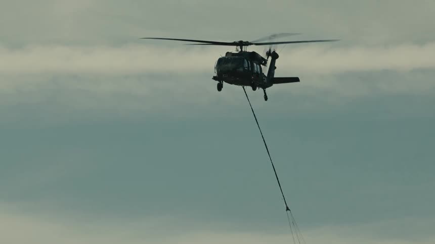 A dark military style helicopter is seen flying against an overcast sky carrying an orange and black bucket suspended below on a long cable for firefighting or external load operations