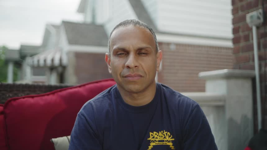 The video is a medium closeup shot of a man with a shaved head and a dark tshirt looking directly into the camera he is sitting on an outdoor patio with red cushions visible and a suburban house faade in the softfocus background