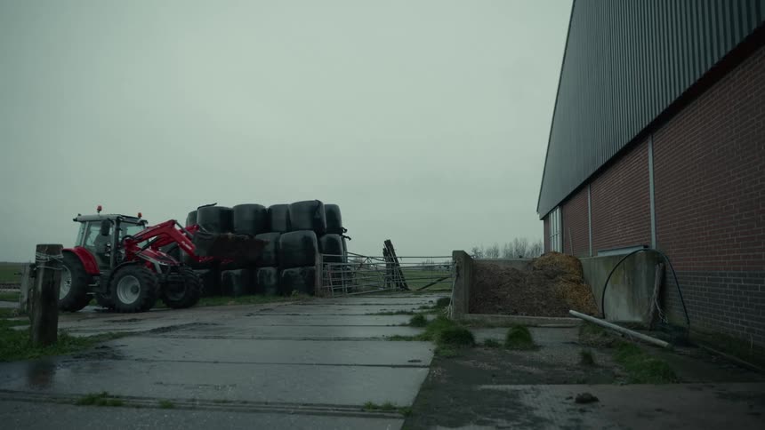 The video shows an exterior slightly gloomy shot of a farm storage area with a large dark metal barn on the right in the foreground there are numerous large round bales of hay wrapped in black plastic stacked on top of each other