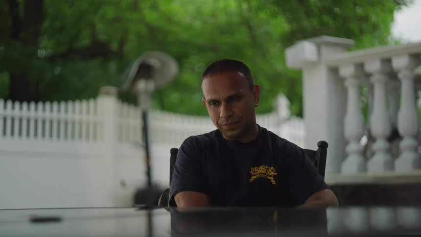 The video shows a man sitting thoughtfully at a dark table on an outdoor patio looking up and making eye contact with the camera the background is a slightly blurred lush green outdoor space framed by white balustrades and a white picket fence