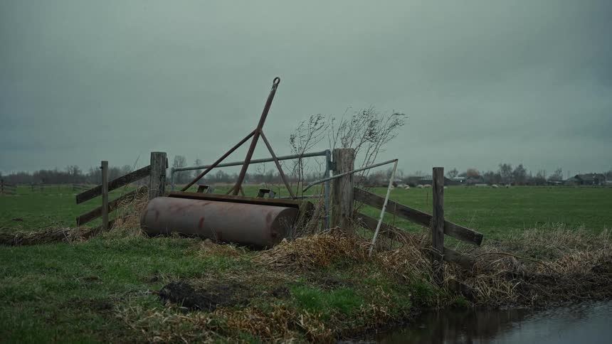 A wide shot shows an old rusted farm roller lying on the ground next to a dilapidated wooden fence and a metal gate with a dark green field and overcast gray sky in the background