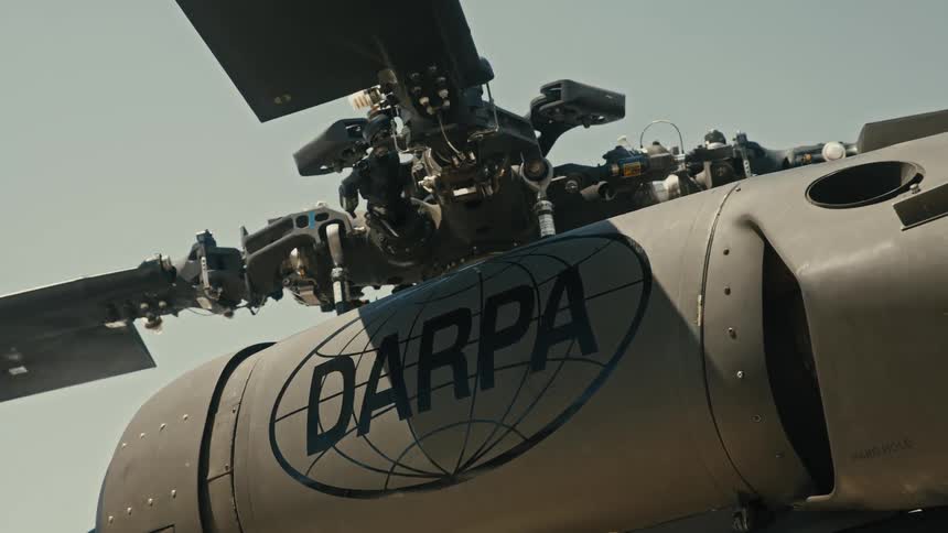 A close angled view shows the rotor head and upper fuselage of a dark military helicopter clearly displaying the darpa logo and text on its side