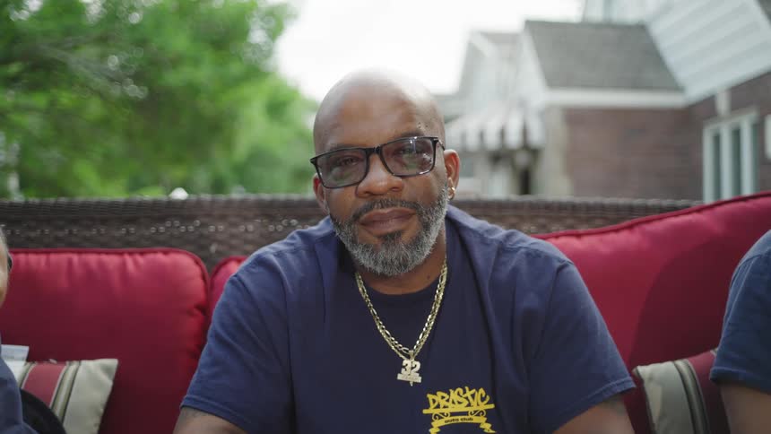 A medium view of an older man with glasses and a gray beard wearing a dark blue tshirt and gold chains as he sits on a red cushion outdoors with a house in the background