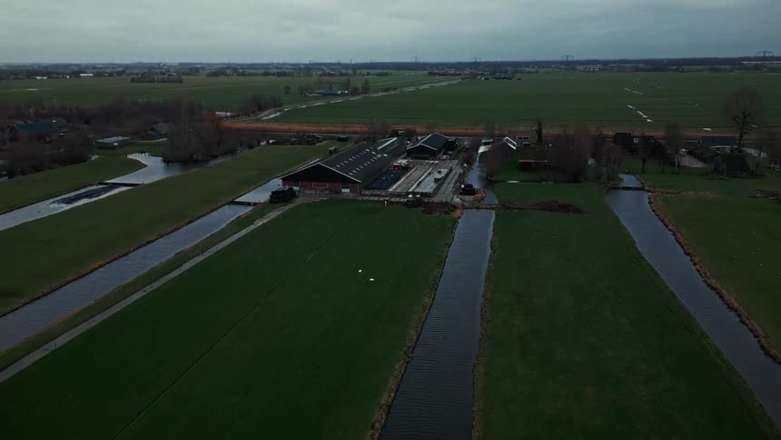 The video is a wide aerial shot of a farm in a flat grassy landscape likely in the netherlands the scene is dominated by long straight irrigation ditches or canals running across the dark green fields towards a cluster of farm buildings