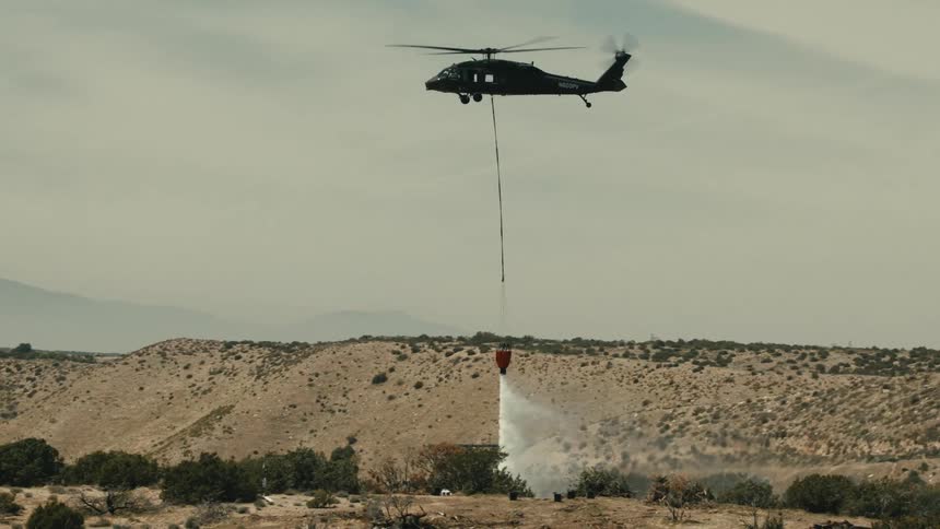 A helicopter watering a desert area from a suspended container during daytime