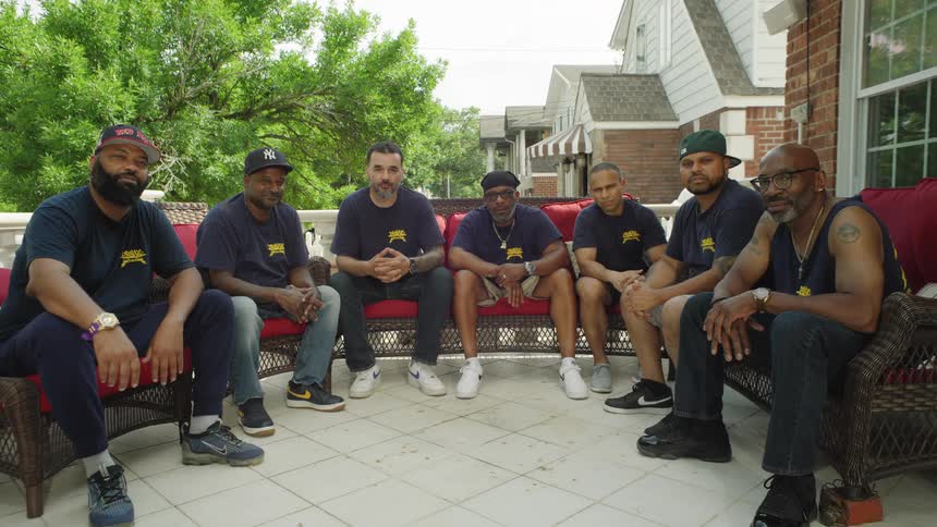 The video shows six men sitting sidebyside on a porch outdoors all wearing dark tshirts with a yellow logo they are facing the camera on a bright sunny day with a suburban house visible in the background