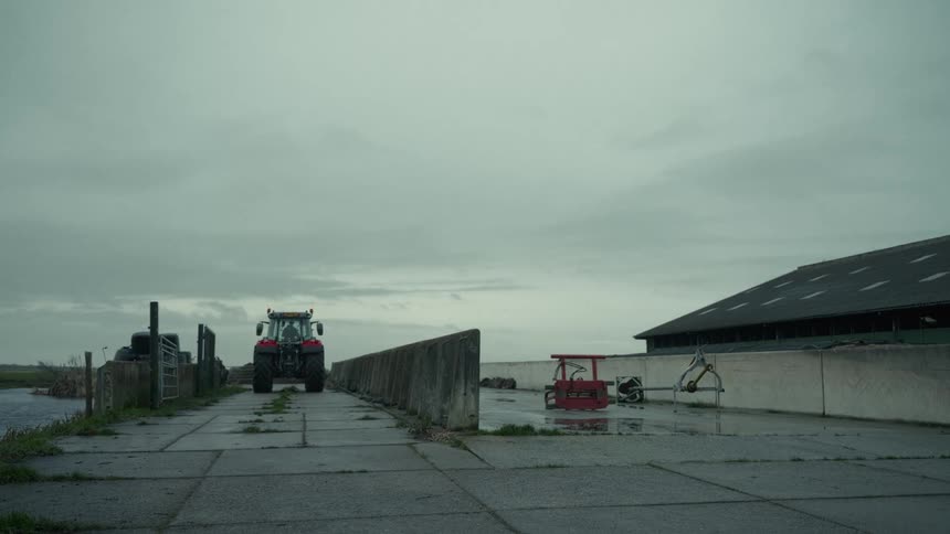 The video shows a lowangle long shot of a red tractor driving from the foreground past the camera and coming to a stop in a paved farmyard area the yard is surrounded by a large dark barn concrete walls and a dull overcast sky