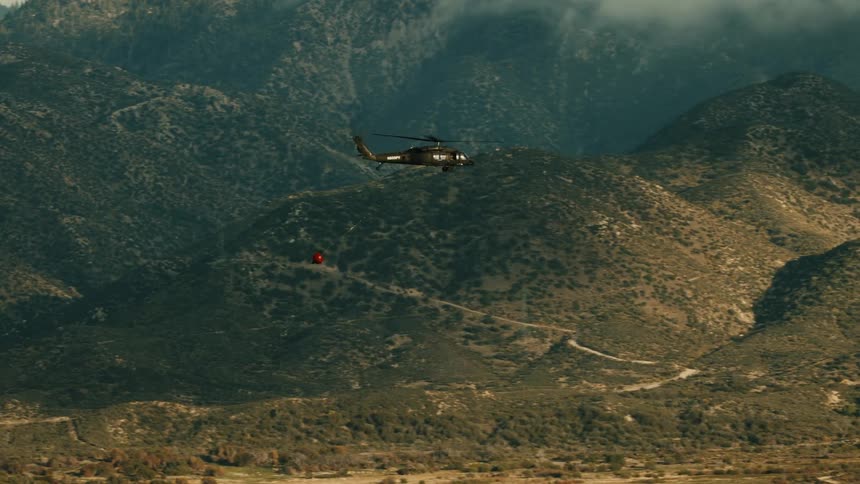 A helicopter flies over a mountain range at midday towing a line with a ball attached beneath it