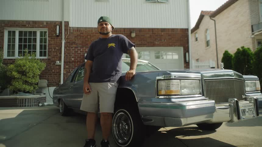 A man leans on a classic lowrider in a suburban driveway