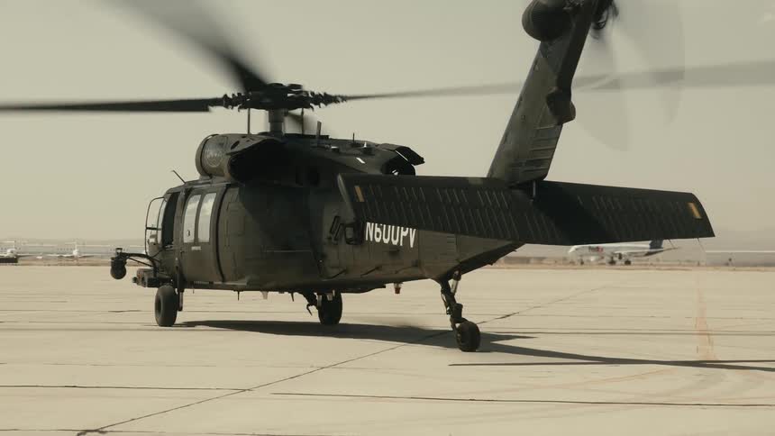 The video shows a darkcolored military helicopter which appears to be a black hawk on an airfield tarmacits main and tail rotors are spinning as it lifts off and begins to move backward kicking up a small cloud of dust