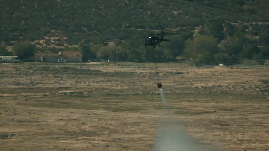 The video shows a helicopter in flight over a wide expanse of dry scrubby terrain likely performing firefighting duties it is seen carrying an orange bambi bucket on a long line which releases a powerful stream of water before the
