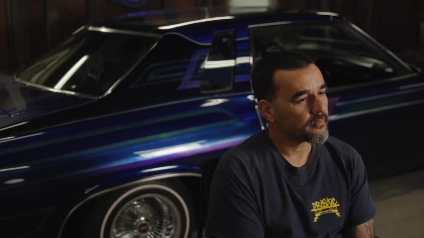 A medium view of a man with a dark beard wearing a dark shirt as he sits thoughtfully in a garage next to a deep purple lowrider car with chrome spoke wheels