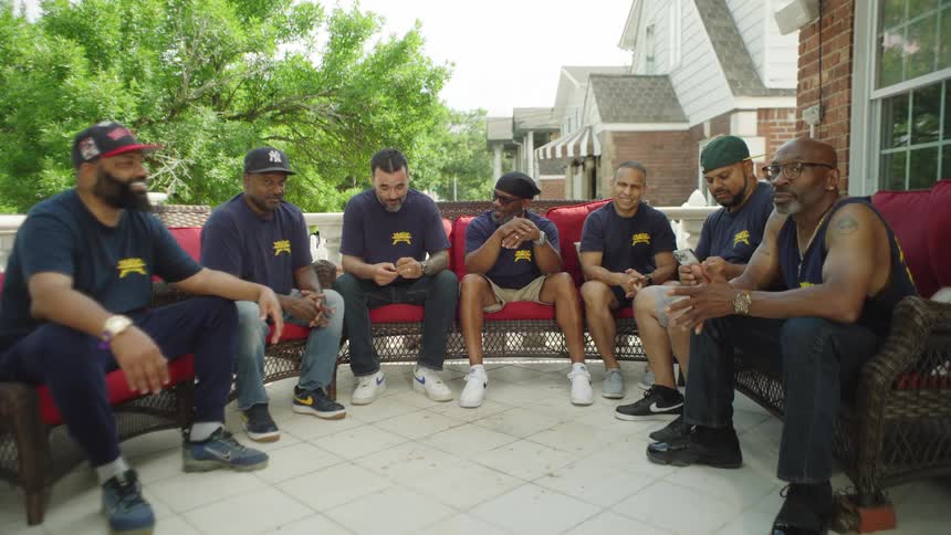 A group of seven men sitting outdoors on a patio they sit in a semicircle in dark chairs all wearing matching dark tshirts and the background shows green trees and a white residential house on a sunny day