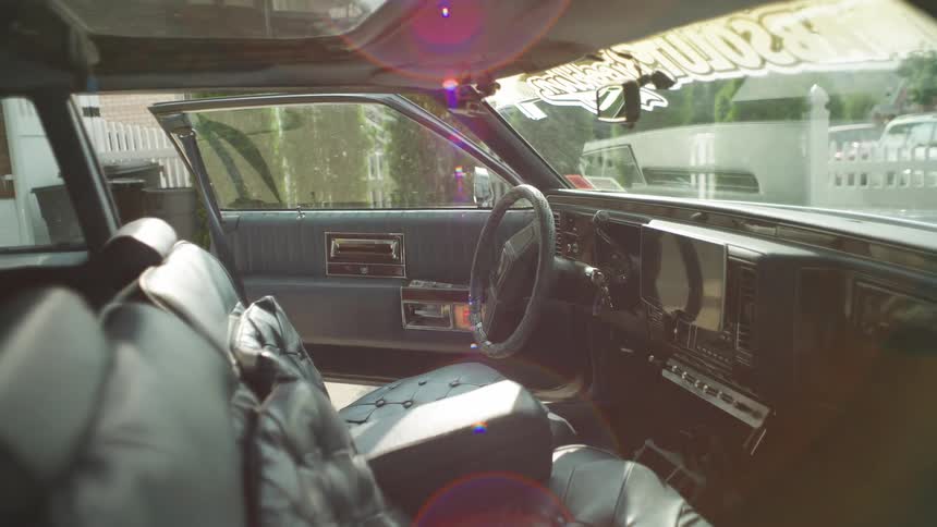 An interior medium view of a vintage american car shows dark blue leather seats and dashboard a sunroof and the passenger side door open in bright sunlight with lens flare