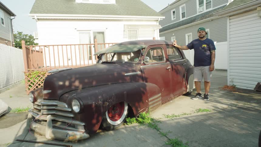The video shows a man standing next to an old heavily rusted darkcolored sedan parked in a driveway in a suburban area the car appears to be a classic model that has been modified featuring lowered suspension and whitewall tires