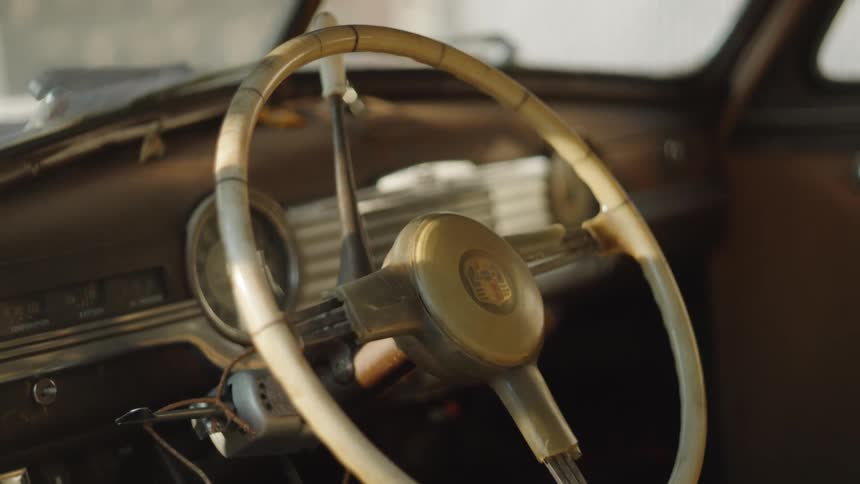 The video is an extreme closeup shot of the dashboard and steering wheel of a classic car with a large offwhite steering wheel dominating the foreground the interior is dimly lit and suggests a vintage vehicle due to the design of the wheel a