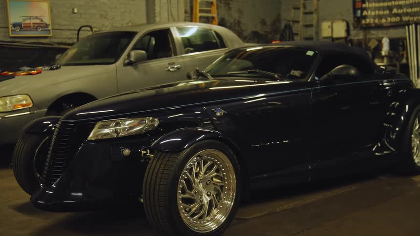 A sleek dark blue plymouth prowler roadster with custom chrome wheels is parked inside a garage or auto shop with a silver sedan parked behind it