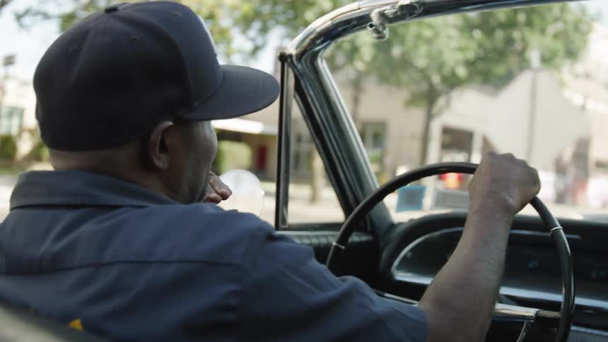 A black man wearing a cap driving a car through the city streets