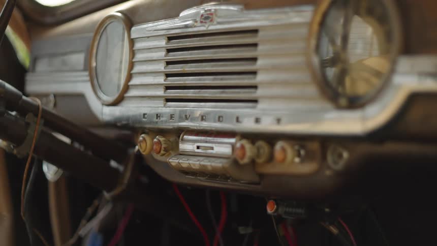 The interior dashboard of an old automobile the dashboard features a prominent chrome grille in the center a circular gauge sits on the right side and various knobs and exposed wiring are visible underneath the main panel