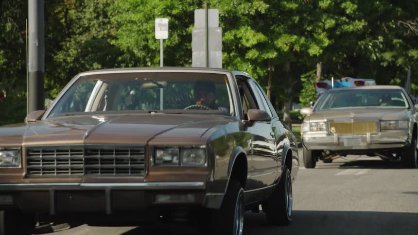 A brown classic coupe car with large wheels drives forward on an urban street a traffic light and other road signs are visible to the left and other vehicles follow in the lane behind the main car under the shade of dense green trees