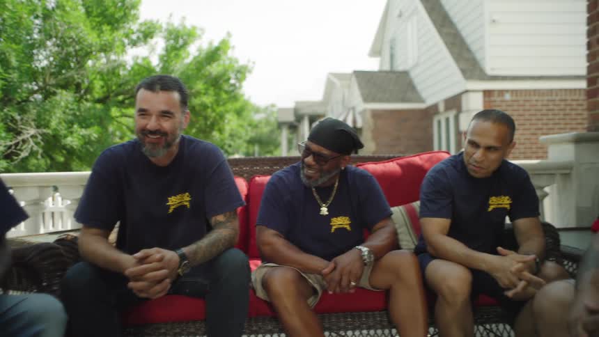 The video is a mediumlong shot of three men sitting on a porch laughing and chatting in an outdoor setting with lush green trees in the background the men are wearing dark tshirts and baseball capsone of which is a new york yankees hatand ap